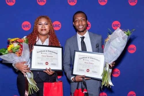 Two students pose in front of a red and blue UIC backdrop holding certificates and bouquets.